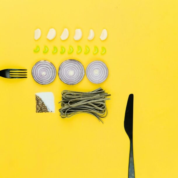 A colorful flatlay of pasta, onion, garlic, herbs, and cutlery on a yellow background.